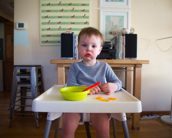 A young toddler with a serious expression sits in a high chair with a yellow bowl and a red spoon in front of them at a wooden table.