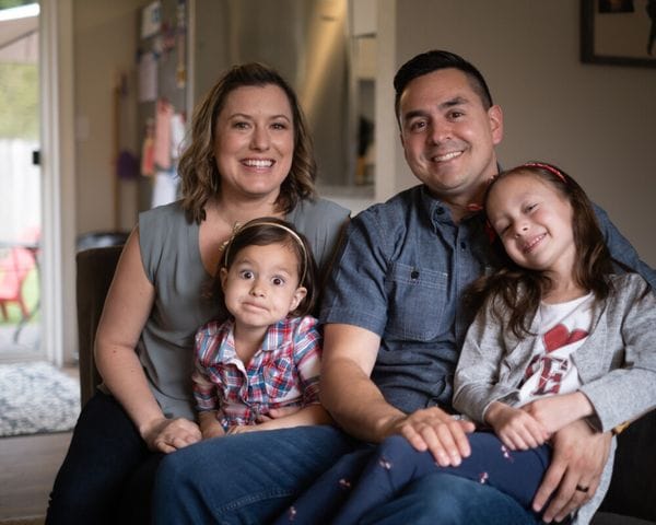 A family of four sits together indoors, with a mother and father smiling behind their two young daughters, one of whom makes a silly face while the other leans against her father.