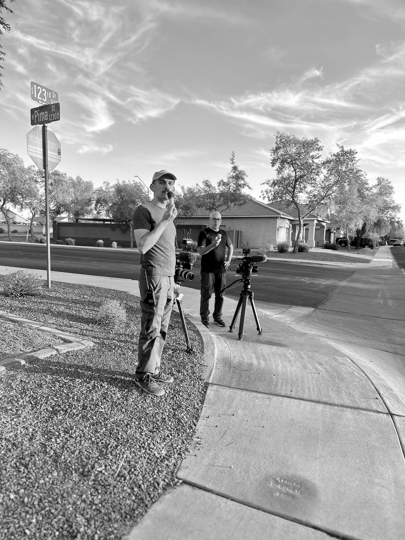 Two men stand on a suburban street corner near the intersection of 123rd Drive and W Pima Street, each holding an ice cream cone while standing next to their professional video cameras mounted on tripods.
