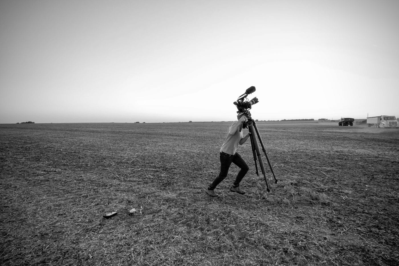 A person in dark jeans and a light shirt leans forward to operate a professional video camera mounted on a tripod in the middle of a vast, empty field, with a drone and its remote lying on the ground nearby.