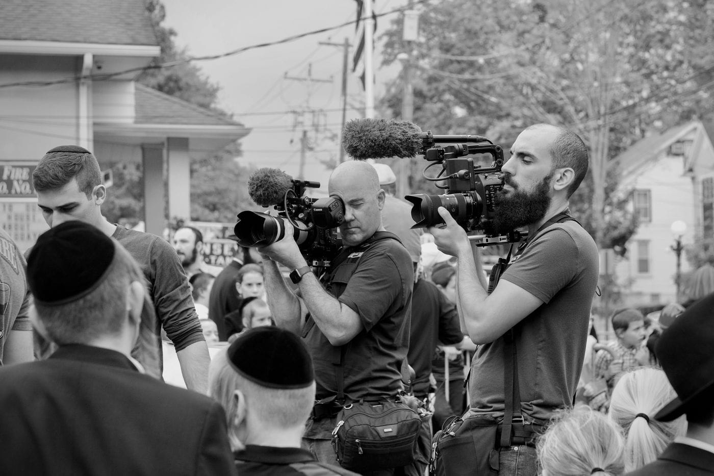 Two men stand side-by-side operating professional video cameras on their shoulders while filming a crowd of people, many of whom are wearing yarmulkes, in a black and white scene.