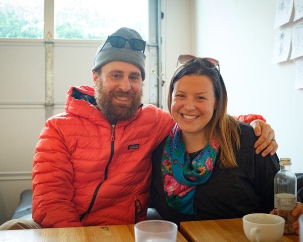 A man in a grey beanie and orange puffer jacket sits with his arm around a smiling woman wearing a colorful floral scarf, both sitting at a wooden table in a brightly lit room.