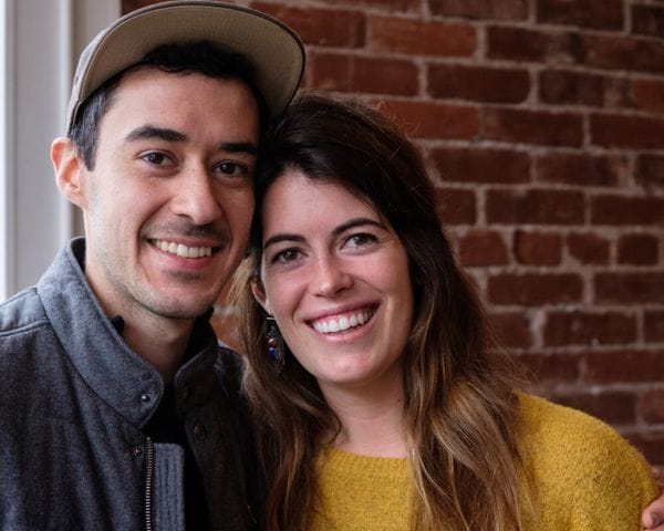 A young man wearing a grey jacket and baseball cap smiles next to a woman with long wavy hair wearing a yellow sweater in front of a brick wall.