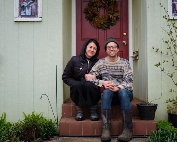 A couple sits together on the steps in front of a red front door with a wreath, the woman wearing a black hooded jacket and the man wearing a patterned sweater, glasses, and a beanie.