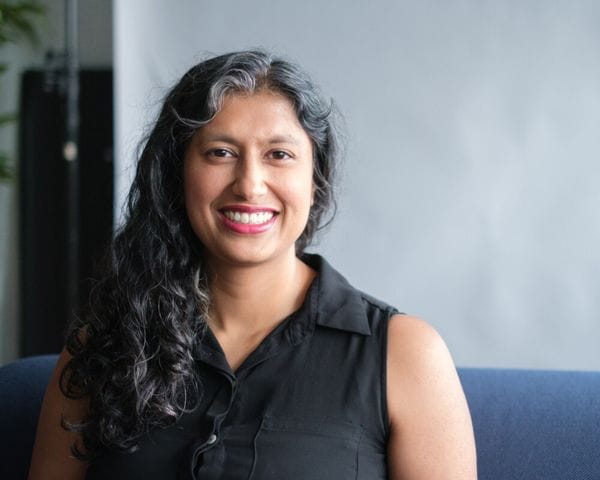 A medium shot of a smiling woman with shoulder-length, curly, dark hair with silver streaks, wearing a sleeveless black button-down shirt and seated against a plain light gray background.