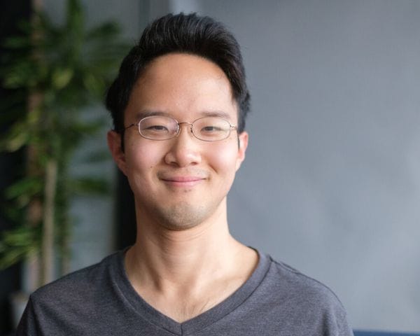 A portrait of an East Asian man with short dark hair wearing wire-rimmed glasses and a gray v-neck t-shirt, posed in front of a blurry background with a plant on the left.