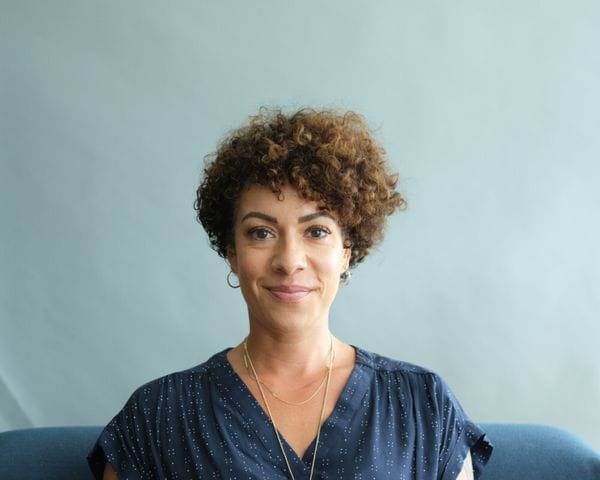 A medium shot features a woman with short, curly brown hair wearing a dark blue, white-patterned top and delicate gold necklaces, centered against a solid, light blue-grey background.