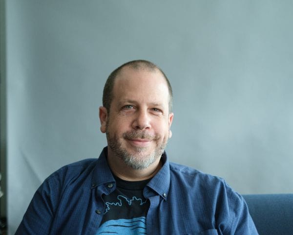A light-skinned man with short hair and a goatee smiles directly at the camera while wearing a blue button-down shirt over a black graphic t-shirt against a light gray background.