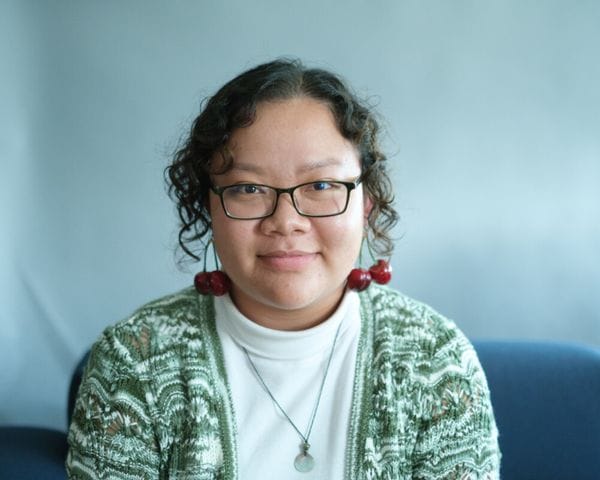 A person with curly dark hair and glasses wears a green patterned cardigan over a white mock-neck shirt, accented by cherry-shaped earrings and a simple pendant necklace.