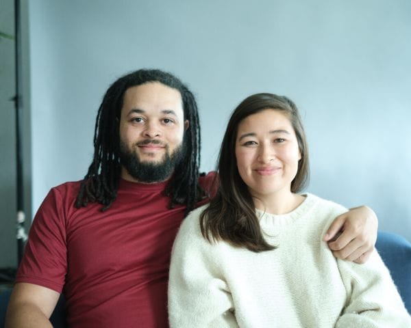 A man with dreadlocks wearing a maroon t-shirt and a woman with long dark hair wearing a white sweater sit together against a light blue backdrop, with the man's arm around her shoulder.