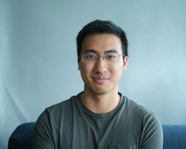 A young East Asian man with short dark hair and rectangular glasses smiles gently while wearing a dark green t-shirt against a plain light blue background.