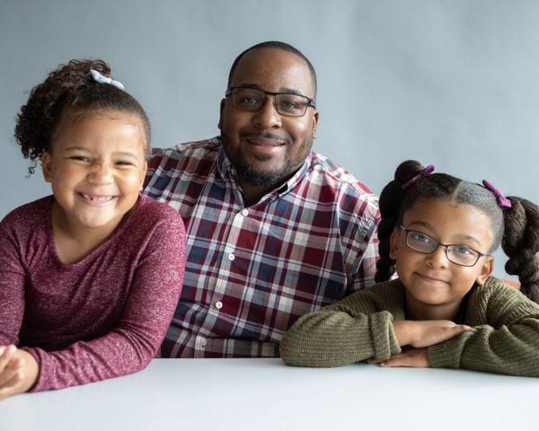 A Black father wearing glasses and a plaid button-down shirt smiles while posing behind a white table with his two young daughters, one wearing a maroon long-sleeve shirt and the other in a green sweater with her hair in two braided pigtails.