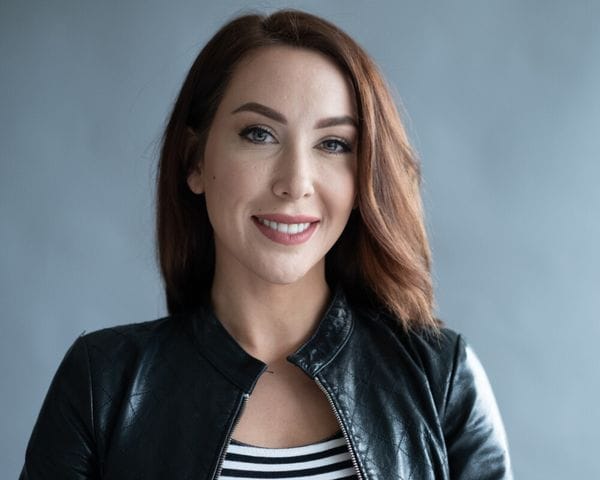 A woman with shoulder-length auburn hair and a friendly smile wears a black leather jacket over a black and white striped top against a plain gray background.