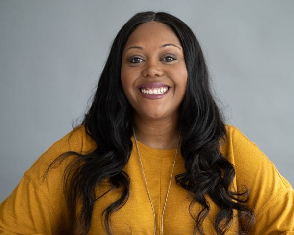 A smiling Black woman with long, dark wavy hair wears a mustard-yellow long-sleeved shirt and a long silver necklace against a plain grey background.