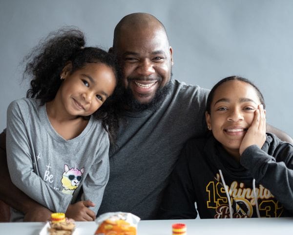 A bald, bearded man smiles warmly while posing for a photo with his two young daughters, who are sitting on either side of him at a table.