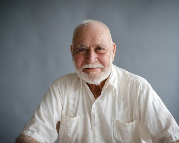 An elderly man with a white beard and short hair wears a white button-down shirt and poses against a plain gray background.