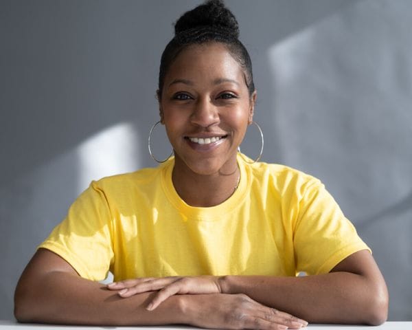 A smiling woman with her hair in a high bun wears a yellow t-shirt and large silver hoop earrings while resting her arms on a table against a gray background.