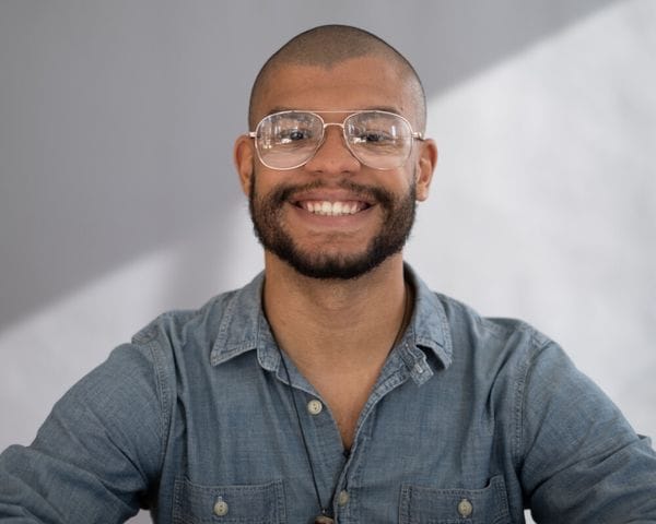 A close-up portrait of a smiling young man with a shaved head and a beard, wearing wire-rimmed aviator glasses and a blue button-down denim shirt, positioned against a light gray, geometric-patterned background.