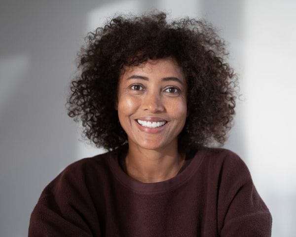 A smiling woman with dark, curly hair and a dark brown sweater looks directly at the camera against a light gray background.