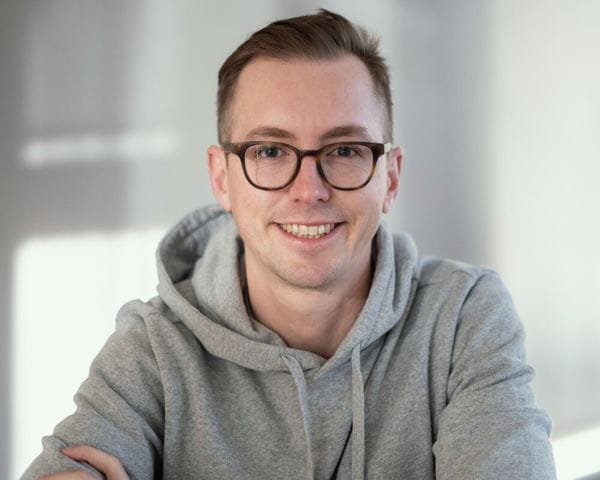 A young man with short light brown hair and glasses smiles while wearing a grey hooded sweatshirt.