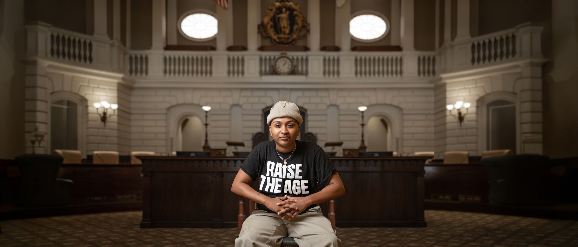 A young person wearing a beige beanie and a black "RAISE THE AGE" t-shirt sits with their hands clasped in front of them in an ornate, wood-paneled courtroom.