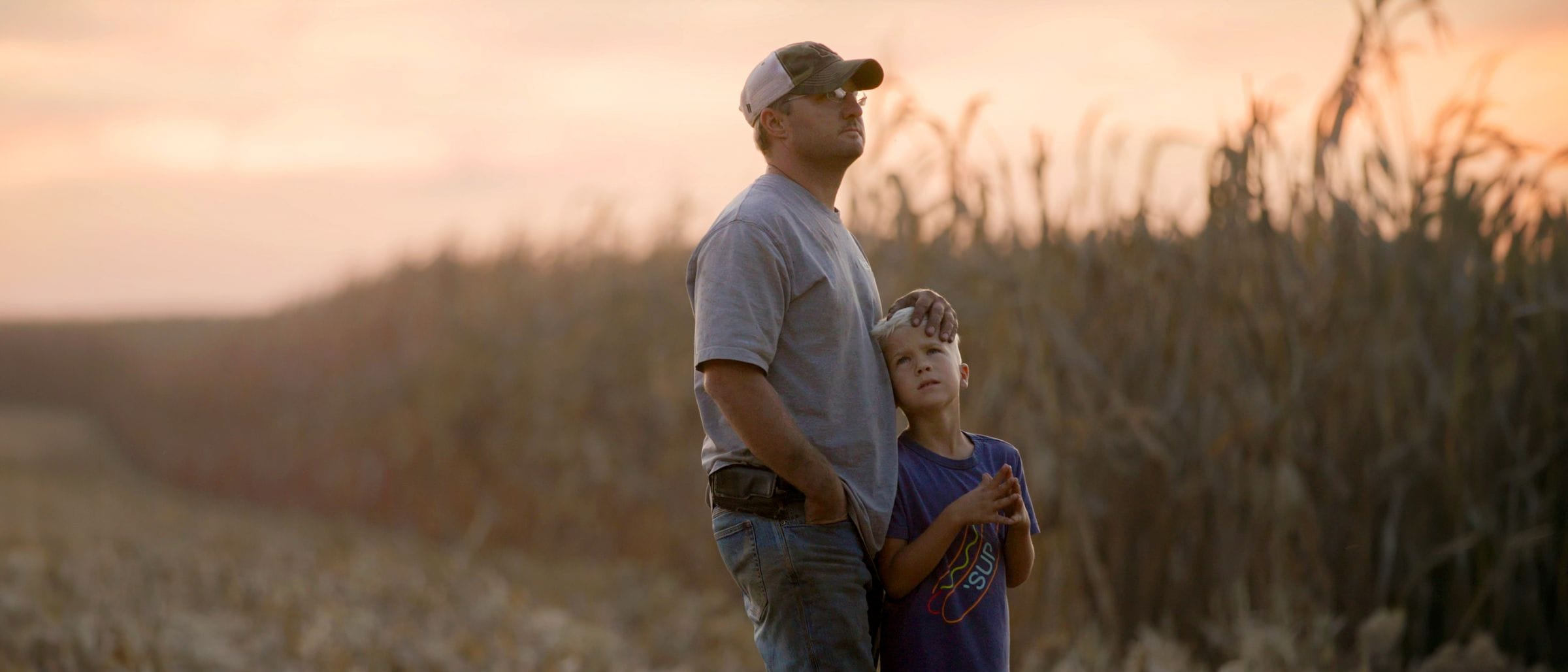 A father wearing a baseball cap and a grey t-shirt stands in a field at sunset with his hand on the head of his young son, who gazes up at him.
