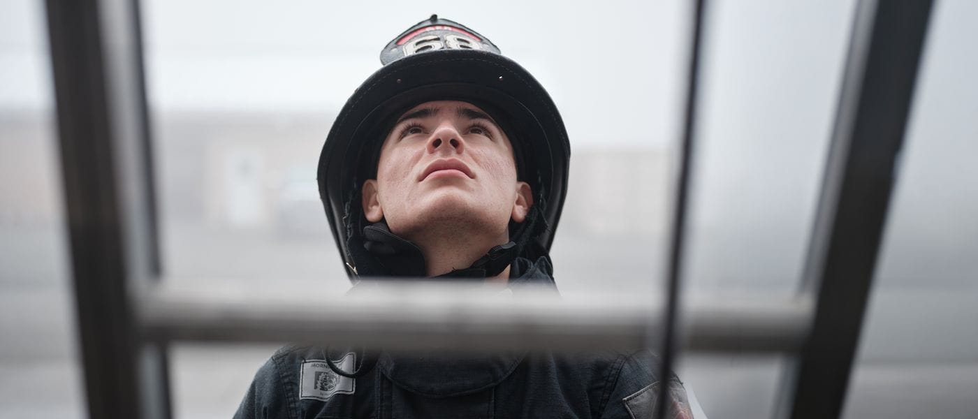 A young firefighter wearing a helmet and turnout gear looks upward while framed by the metal rungs of a ladder.