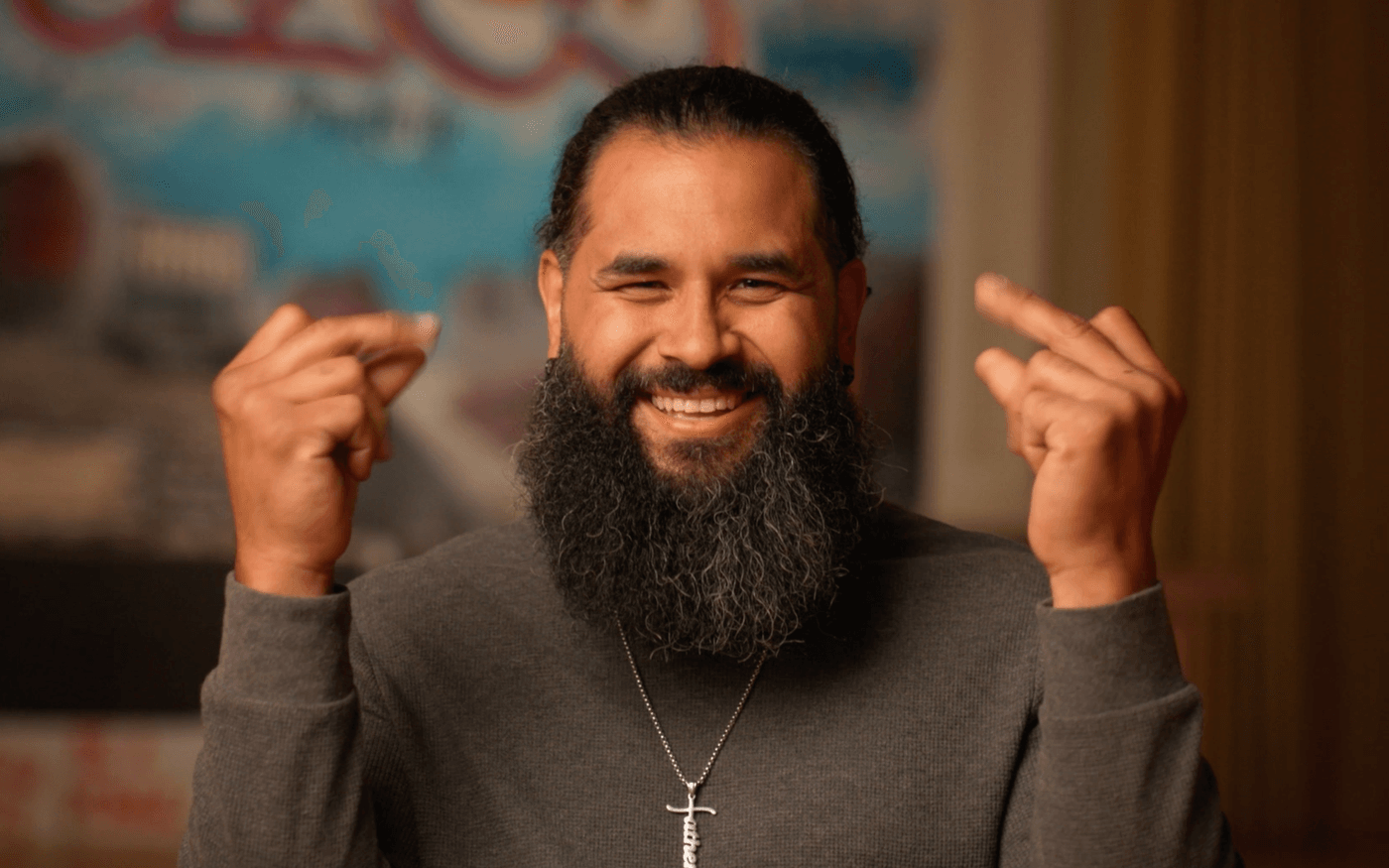 A smiling man with a full beard and dark hair tied back wears a grey long-sleeved shirt and a cross necklace while holding both hands up near his head with his middle fingers raised.