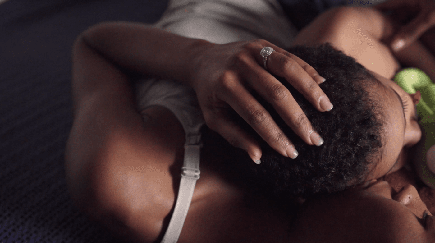A person with dark skin wears a sparkling diamond ring as they tenderly rest their hand on the head of another person whose face is partially visible against their chest.