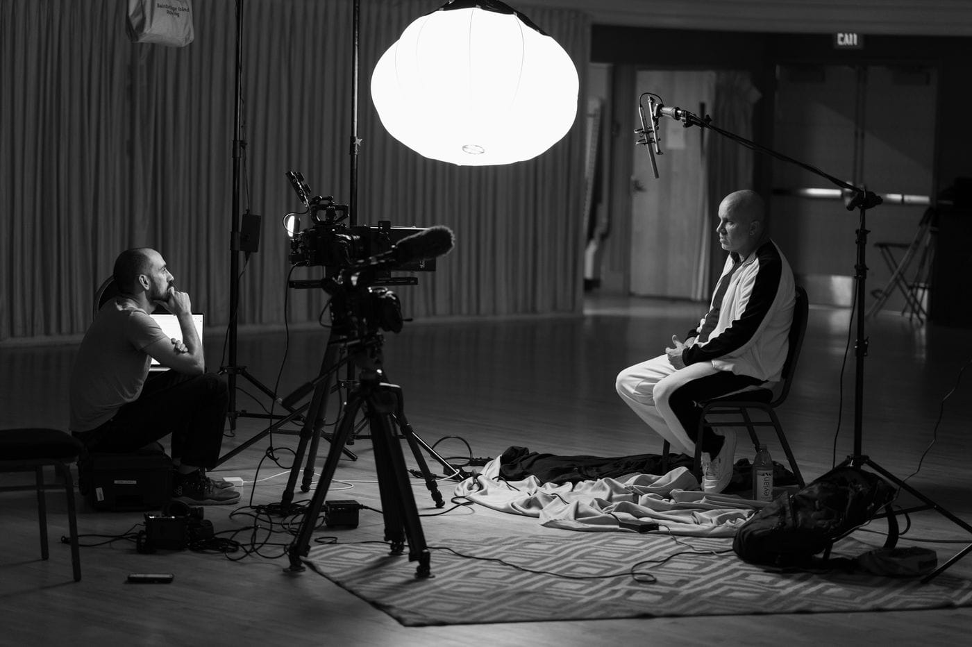 A film crew member sits on the floor operating a camera and laptop while filming a bald man wearing a tracksuit who sits in a chair under a large, glowing light sphere in a studio setting.