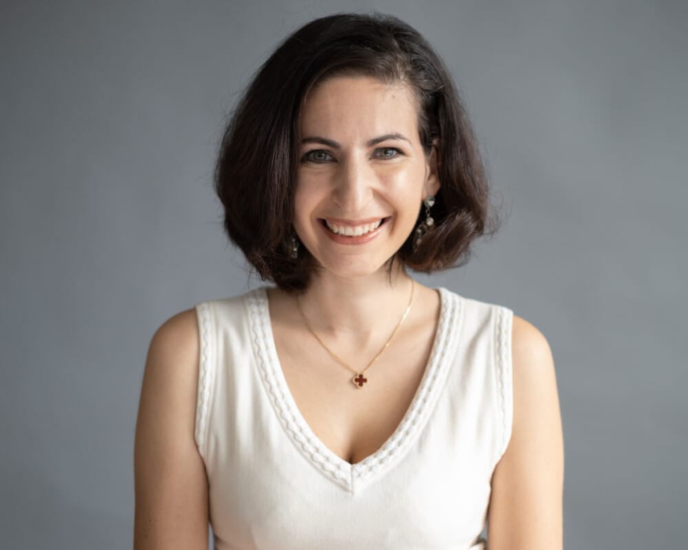 A smiling woman with dark, shoulder-length hair wears a white sleeveless V-neck top and a small clover-shaped necklace against a plain gray background.