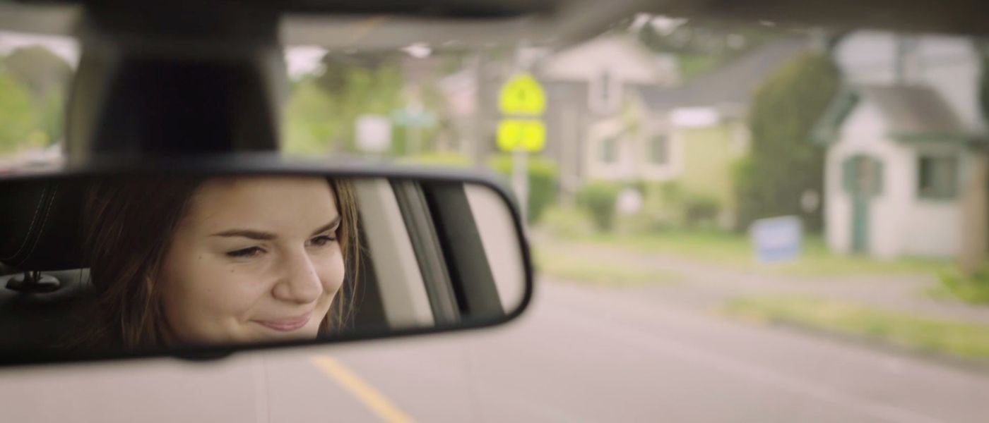 A young woman with brown hair smiles softly as seen through a car's rearview mirror while driving down a residential street.
