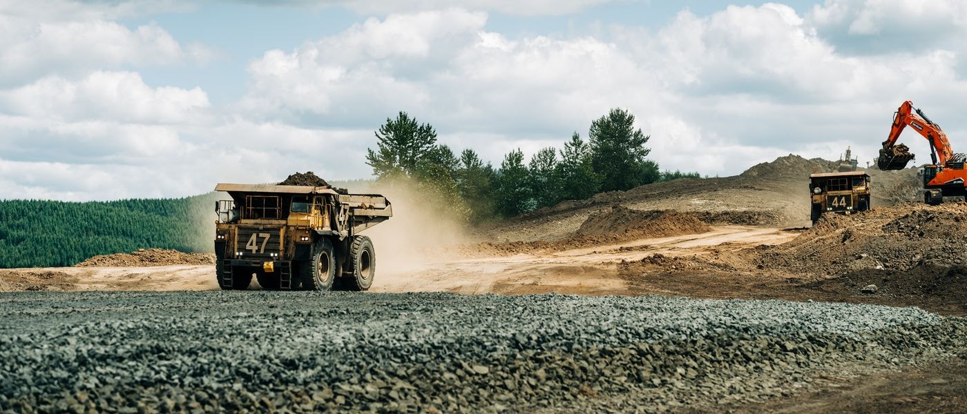 Two large yellow mining dump trucks, numbered 47 and 44, operate on a dusty dirt road near a pile of gravel while an orange excavator loads earth in the background under a cloudy sky.