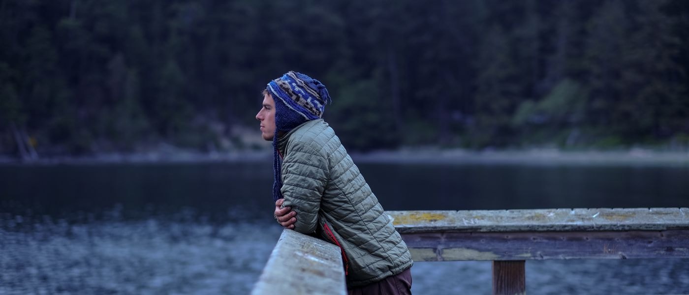A young man in a blue patterned knit hat and olive green puffer jacket leans against a weathered wooden railing, looking out over a calm, dark lake with a forested shoreline in the background.