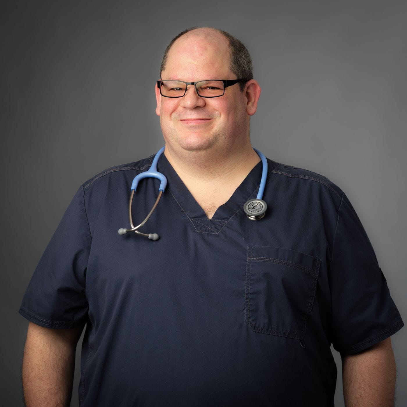 A middle-aged man with a receding hairline and glasses wears dark blue medical scrubs and a light blue stethoscope around his neck while standing against a gray backdrop.