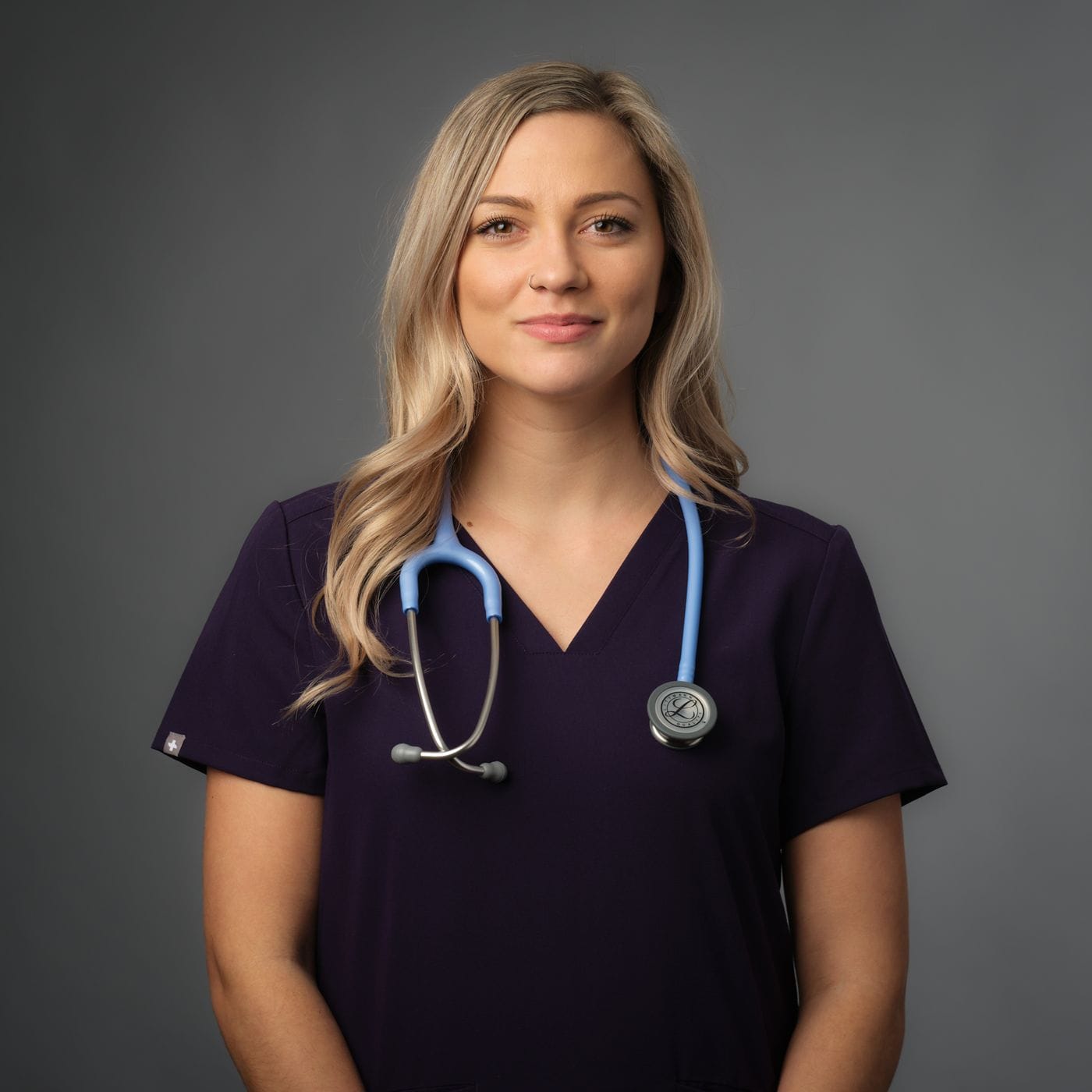 A blonde woman with a nose piercing wearing dark purple medical scrubs and a light blue stethoscope around her neck stands against a solid gray background.