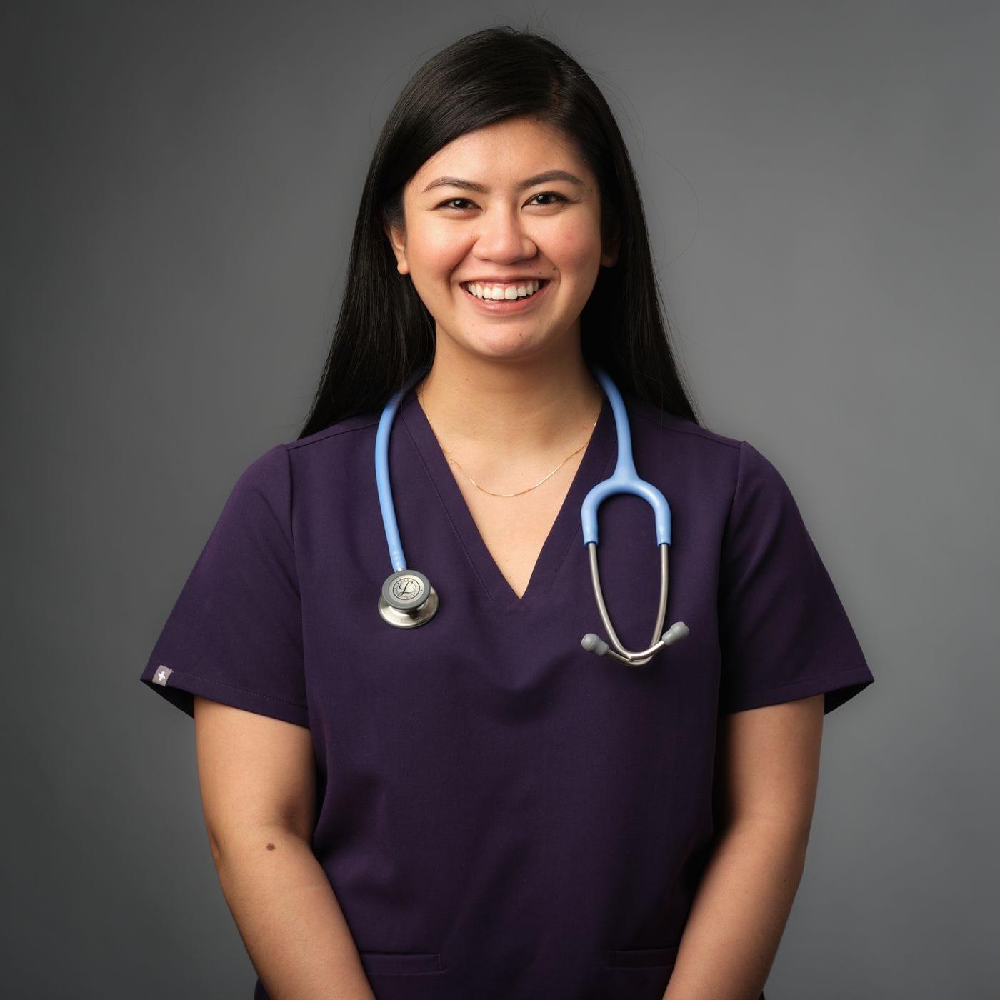 A smiling woman with long dark hair wears a purple scrub top and a light blue stethoscope draped around her neck against a solid gray background.