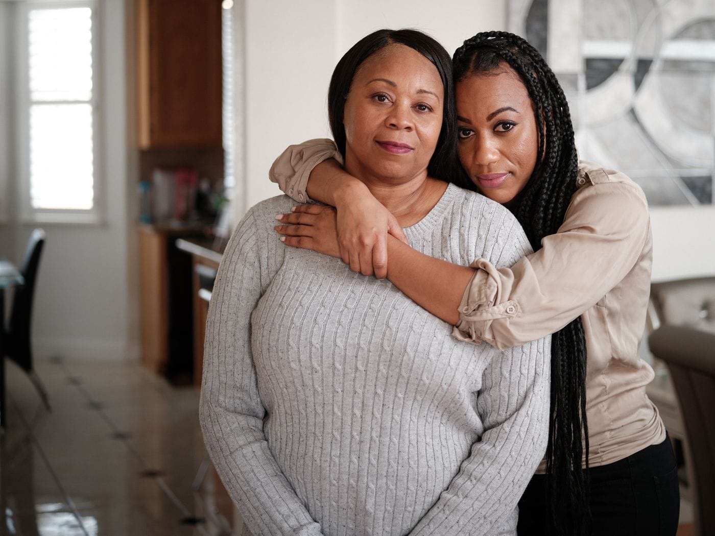 A younger Black woman with long braided hair stands behind an older Black woman in a gray sweater, resting her chin on the older woman's head while embracing her from behind.