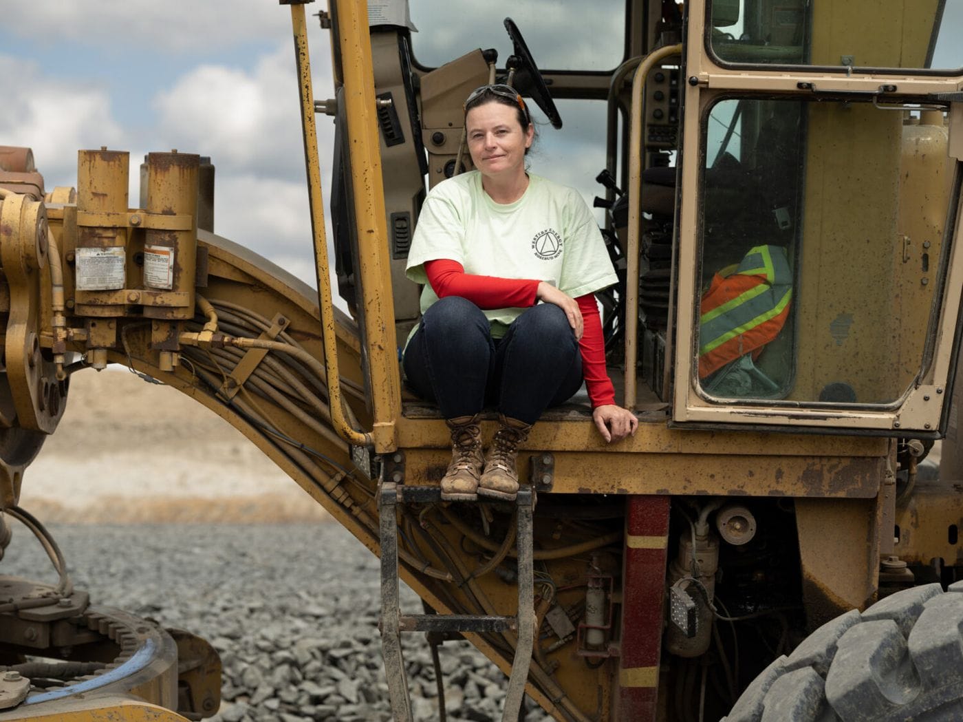 A woman in a light-colored t-shirt and red long-sleeved shirt sits on the steps of a large yellow construction vehicle at a worksite.