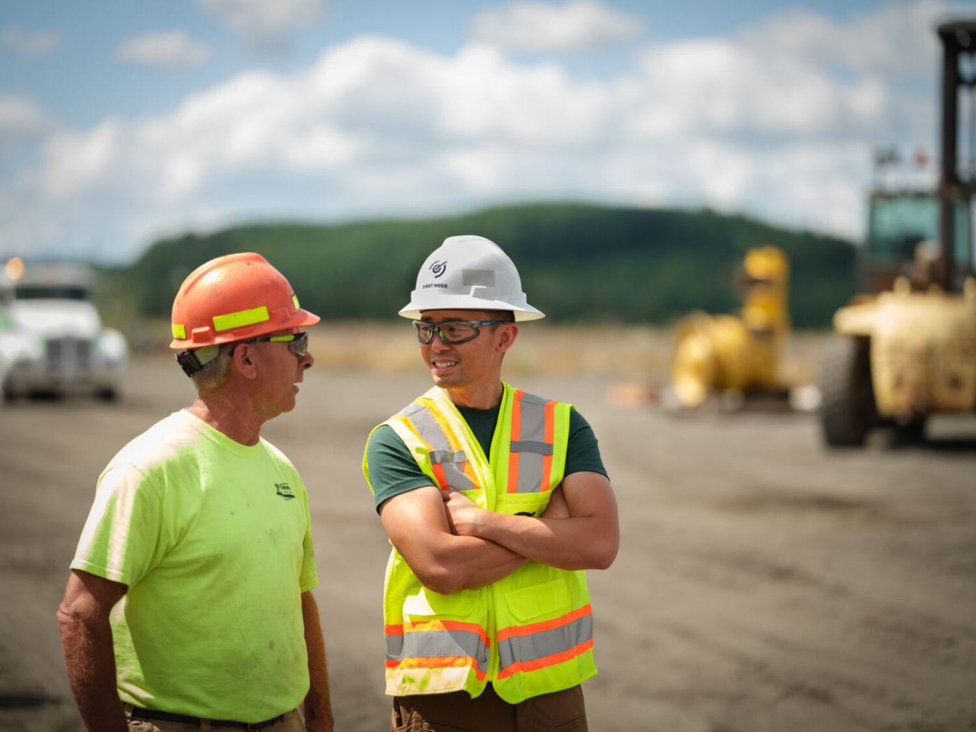 Two construction workers in safety gear and hard hats stand on a dirt lot, engaged in conversation with heavy machinery visible in the blurred background.