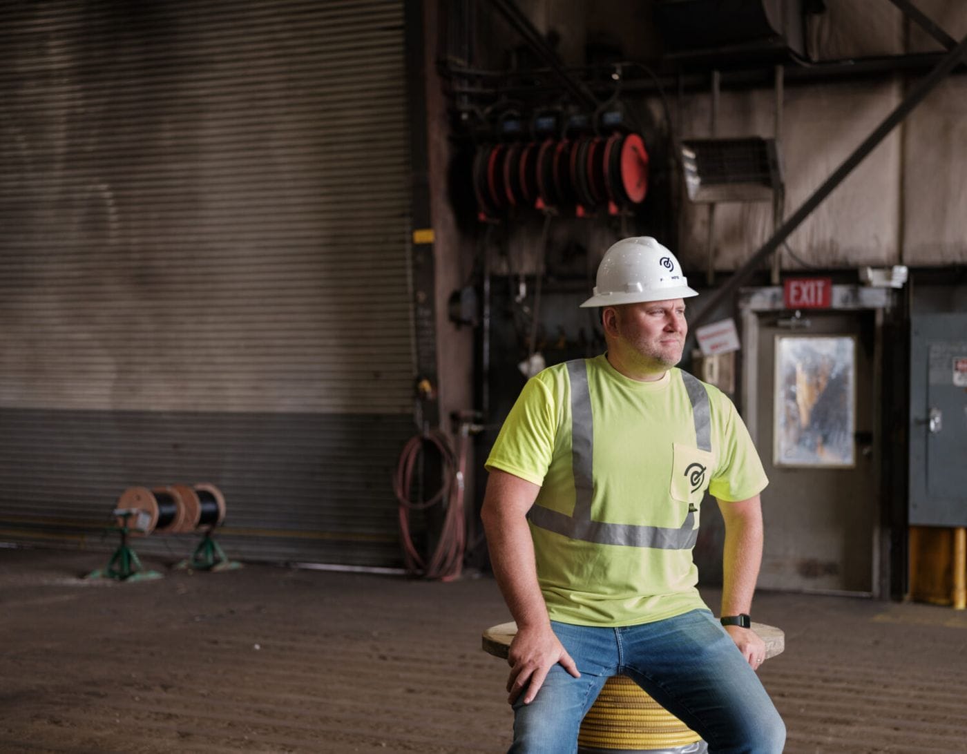 A man wearing a white hard hat and a high-visibility yellow t-shirt with a safety vest sits on a yellow industrial spool inside a large warehouse facility.