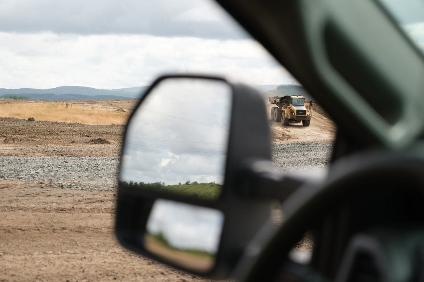 A view from inside a vehicle looking through the side-view mirror at a large yellow dump truck driving on a dusty, unpaved construction site under a cloudy sky.