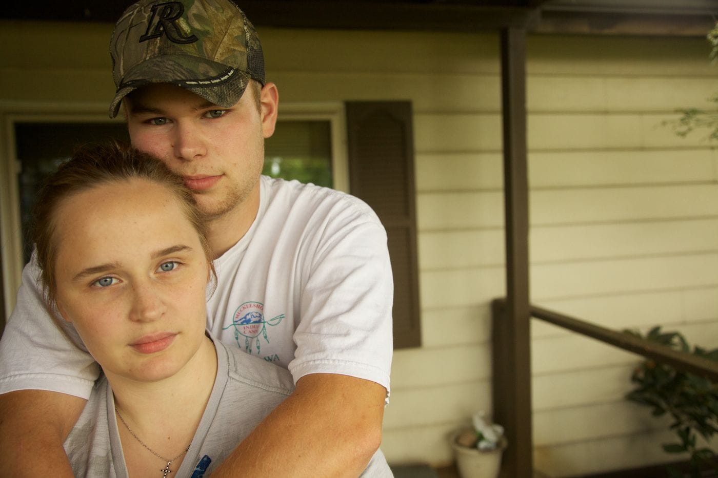 A young man wearing a camouflage baseball cap stands behind and embraces a young woman in front of the siding of a house.