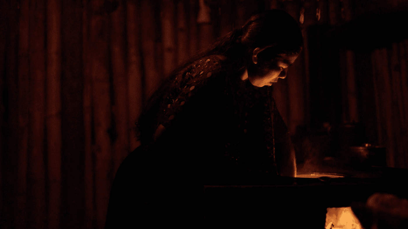 A woman with long hair wearing a textured patterned top stands in a dim room with bamboo-slatted walls, leaning over a glowing, steam-filled cooking area illuminated by a fire below.