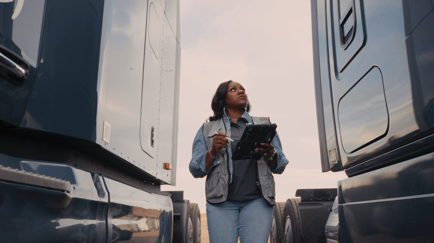 A woman in a grey vest and denim jacket stands between two large semi-trucks, looking upward while holding a digital tablet and stylus.