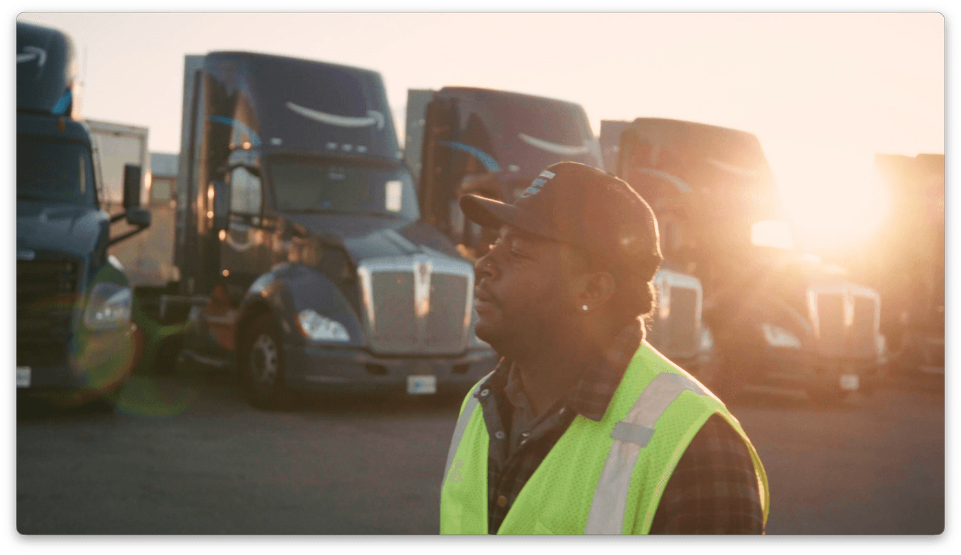 A man wearing a high-visibility yellow safety vest and a cap stands in front of several parked Amazon semi-trucks at sunrise.