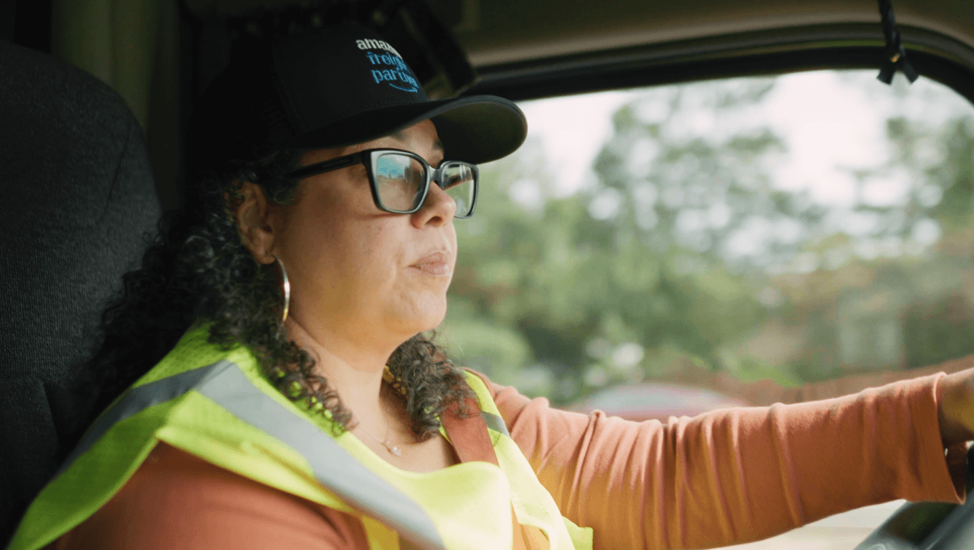 A woman with curly dark hair wearing glasses, a high-visibility yellow vest, and an Amazon Freight Partner cap sits in the driver's seat of a truck, focused on the road ahead.