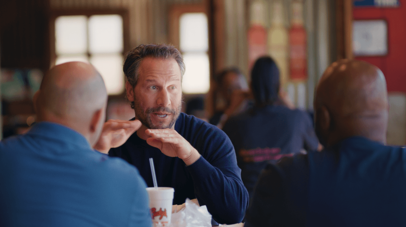 A man with graying hair and a beard sits at a table and talks to two other men, gesturing with his hands while a disposable cup from Rudy's sits in front of him.