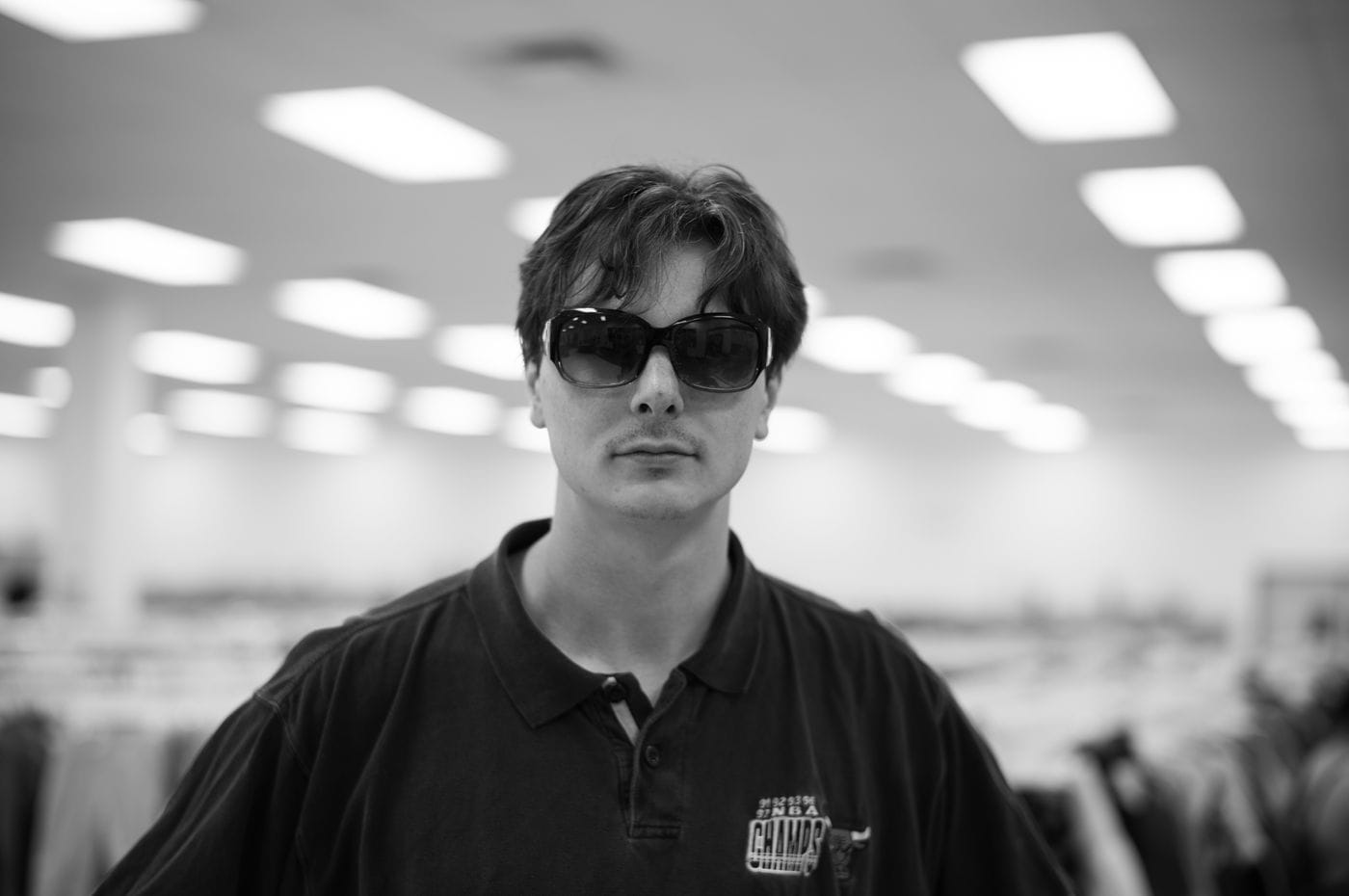 A black-and-white, eye-level portrait shows a young man with dark, slightly unkempt hair wearing dark sunglasses and a polo shirt with a graphic print, standing in a brightly lit, out-of-focus retail store.