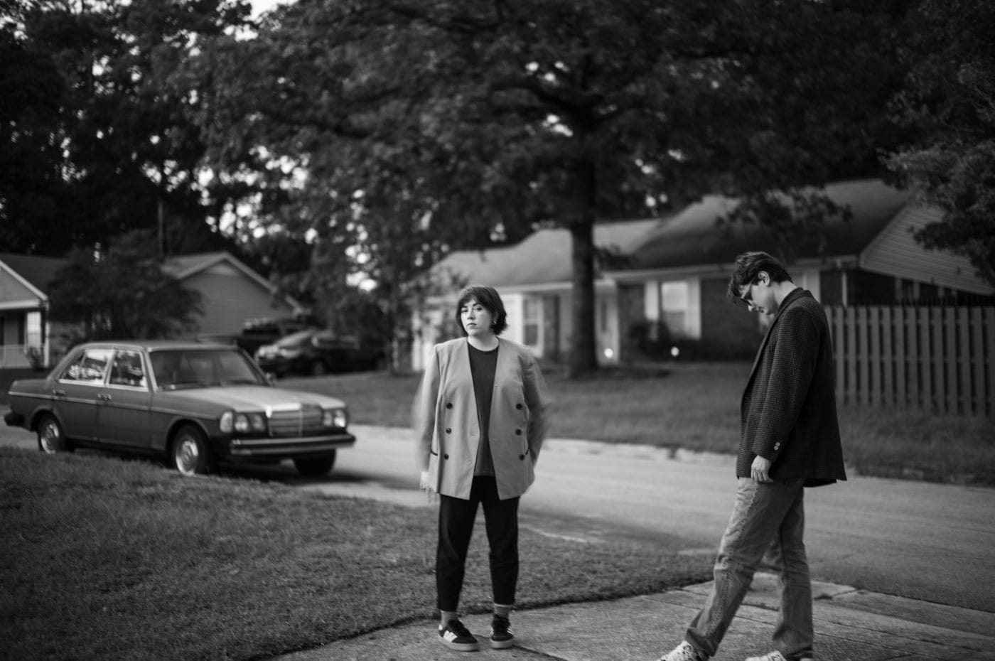 A young woman in a blazer stands looking at the camera on a suburban sidewalk while a young man in a jacket and jeans walks beside her, captured in a black and white photograph with a motion-blurred effect.